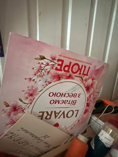 The image shows a close-up of several items stacked together, possibly in a storage area or a cluttered shelf. The main subject is a book or magazine with a pink cover adorned with cherry blossoms. The text on the cover is in Ukrainian. Large red letters spell out "ПОРЕ" vertically on the left side. To the right, within a circular emblem, are the words "Вітаємо з весною" (Greetings with spring) and below that, "LOVARE". A handwritten note is attached to the cover, with visible text including "1410182" and what appears to be a date "14/10/18".

Other objects in the background are out of focus but suggest a collection of assorted items. These include what looks like spools of thread, possibly orange and white, a dark blue object, and a metal wire handle, possibly from a tool or a container. The background appears to be a ribbed surface, like corrugated metal or a slatted shelf.

The lighting is somewhat dim, and the overall impression is one of a candid snapshot rather than a posed photograph. There are no people visible. The setting does not provide clear external location cues, but the handwritten note and the text on the book suggest a Ukrainian context, and the provided location context is Podilsk, Ukraine. The content of the book or magazine seems to be spring-themed.