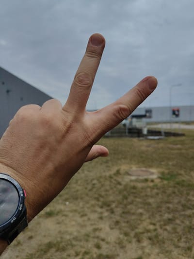 The image shows a person's hand making a peace sign with their index and middle fingers extended, against a backdrop of an overcast sky and dry, brown grass. In the background, a large, flat-roofed building with a gray facade and some indistinct signs is visible. The overall scene appears to be outdoors, possibly in an industrial or commercial area given the building's architecture. The weather seems to be cloudy and dull, suggesting it could be morning or late afternoon. The person is wearing a black watch with a round face on their wrist. The image conveys a casual and possibly hopeful gesture of peace.