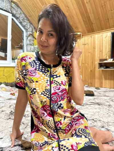 A woman with dark hair is sitting on a patterned carpet in a room with wood-paneled walls and a wooden ceiling. She is wearing a short-sleeved zippered dress with a colorful floral and polka dot pattern. She is looking towards the camera with a slight smile and has her right hand raised, touching her hair. To the left of the woman, a window is visible with a white frame, and beyond it, a textured wall with stone-like accents. A white plastic bag with some items inside is placed on the carpet near the window. In the background, there is a shelf unit with some objects on it. The environment appears to be indoors, possibly a residential space.