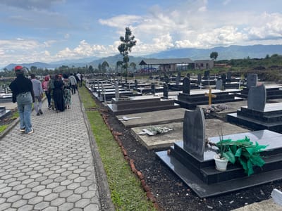 The image shows a cemetery located at -1.6 latitude and 29.124 longitude in Nzulo, Democratic Republic of Congo. There are numerous black granite tombstones with wooden crosses in the cemetery.  People are walking on a stone pathway away from the camera toward a small, white building in the distance.  The cemetery is surrounded by green grass, and the background contains a blue sky with white clouds.  There are several mountains in the distance.