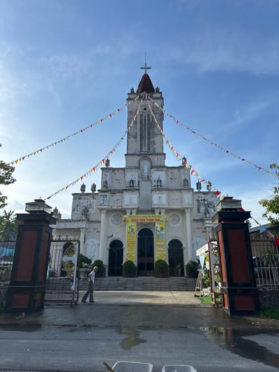 A wide-angle, eye-level shot captures the facade of a church in Diên Điền, Vietnam, bathed in daylight under a bright blue sky with scattered clouds. The church, painted a faded white with grey accents, features a tall, prominent bell tower topped with a cross. A string of colorful triangular flags stretches across the frame, connecting the church to poles on either side of the gate. The entrance to the church is marked by a wide set of stairs leading up to large arched doorways. Two banners hang on either side of the central entrance, one with text and an image of a religious figure, and another with Vietnamese text.
In the foreground, a paved road with puddles of water stretches towards the church, indicating recent rain or washing. A man in grey pants and a white shirt walks away from the camera towards the church gate. He is partially obscured by a large, dark-colored pillar that forms part of the gate structure. The gate itself is ornate and made of dark metal with gold accents. A second person, appearing to be a child, is visible behind the gate. The environment is tropical, with visible greenery and trees to the left and right of the church. The overall scene suggests a community event or celebration given the decorative flags.