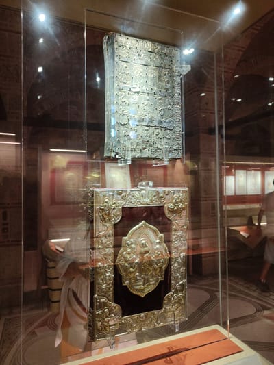 In the city of Kalambaka, Greece, within a museum exhibit, two ornate, ancient books are displayed in glass cases. The upper book, likely a relic or manuscript, is covered in intricate, metallic relief work, featuring rows of figures and decorative patterns. It rests on a clear acrylic stand. Below it, a second, larger book is encased. Its cover is adorned with a deep red velvet backing, embellished with elaborate silver or gold metalwork. The central focus of this lower book's cover is a detailed bas-relief depicting figures, possibly religious in nature, surrounded by ornate borders and corner decorations. The exhibit space appears to be dimly lit, with spotlights illuminating the artifacts. In the background, through the glass, other displays and museum architecture are visible, including what appear to be framed documents and arches. A person, partially obscured by reflection and distance, is seen in the far background, walking away from the camera.