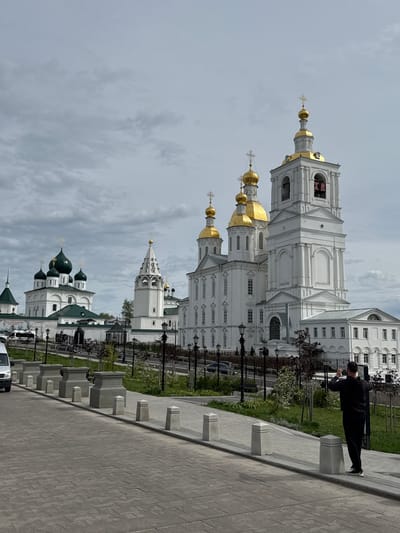 The image depicts the Cathedral Square in Arzamas, Russia, showing multiple churches with white walls and green and gold domes. A man in a black jacket and pants is standing on a low concrete wall, appearing to take a picture with his phone. The foreground features a paved walkway with concrete bollards and grass. Street lamps are visible along the walkway. The sky is overcast.