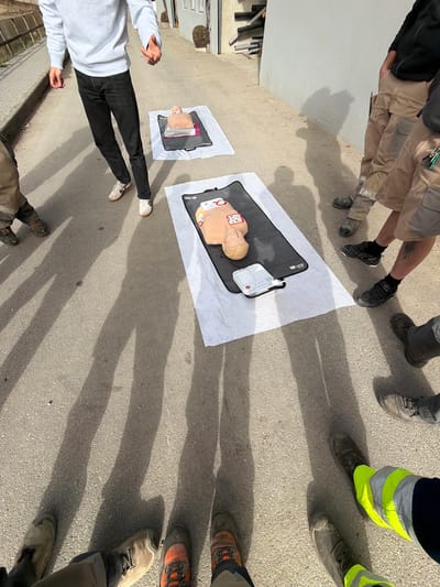 The scene depicts an outdoor first-aid or CPR training session occurring on a paved surface in Adliswil, Switzerland. Two medical training manikins are centrally positioned on black mats, which are laid over white towels on the ground. The manikin in the foreground is an adult-sized torso, light-skinned, with chest compression pads affixed to its sternum and a white electronic CPR feedback device or AED trainer situated at its feet, connected. A second, smaller manikin, likely representing a child or infant, is placed further back on a similar setup.
Multiple individuals are present. One person, visible from the waist down in a white sweatshirt, black pants, and white sneakers, stands on the left, gesturing with their hands. Other individuals are partially visible on the right and in the immediate foreground, primarily showing their lower bodies. They wear varied attire including khaki shorts and trousers, work boots, and, in the foreground, reflective safety yellow legwear. Elongated shadows of the individuals are cast across the asphalt or concrete surface, indicative of direct sunlight. The background consists of plain, light-colored building walls and a stack of pipes or conduits against one wall.