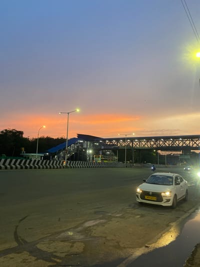 The image depicts a street scene at dusk in the St. Thomas Mount-Pallavaram Cantonment Board, India. A white sedan with license plate TN05CW5223 is parked on the right side of the road, its headlights illuminated. The road surface appears wet, with reflections from streetlights and the sky. In the background, a pedestrian overpass with blue roofing spans across the road, illuminated by overhead lights. A bus is partially visible on the left side of the overpass. The sky displays a gradient of colors, transitioning from blue at the top to orange and pink hues near the horizon, indicating sunset. Streetlights are visible along the road and on the overpass. Trees line the background beyond the overpass. The scene captures a moment of transition from day to night in an urban environment.