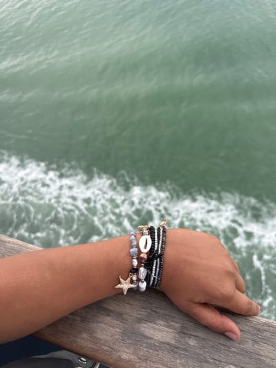 A top-down medium shot shows a person’s forearm and hand resting on a weathered wooden surface, likely a pier or boardwalk, with a view of the ocean in the background. The person is wearing multiple bracelets, including beaded ones with a seashell and a starfish charm. The ocean water is a greenish-blue hue, and there are visible whitecaps, indicating slight turbulence. It is daytime, and the lighting is soft. The location is likely Loon-Plage, France, given the ocean view.
