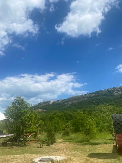The image depicts an outdoor scene with a clear blue sky dotted with white clouds. In the foreground, there is dry grass and a circular concrete structure with a dark lid. To the left, there is a wooden picnic table and grill, and a white tent structure. Further into the scene, there are several trees and dense green foliage, leading to a mountainous landscape in the background. A small wooden structure with a dark roof is visible on the right side of the image. The scene appears to be a rural or natural setting, possibly a campsite or park, within the vicinity of Орлиновский округ, Ukraine. There are no people or animals visible in the image.