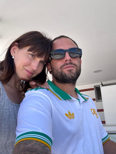 A selfie depicts a man and a woman positioned close together, possibly on a boat, though the location is not explicitly specified as Pomezia, Italy. The woman is in the foreground, wearing a grey shirt and a delicate necklace, with short brown hair featuring bangs. She has light eyes and an earring on one ear. Behind her, a man with a beard, a nose ring, and sunglasses rests his face near hers. He is wearing a white soccer jersey with green and gold trim and an Adidas logo. His arm is visible, showing tattoos. The background consists of a white wall, likely a part of the boat's structure, with a small speaker and a rectangular window visible. The lighting appears to be bright and natural.