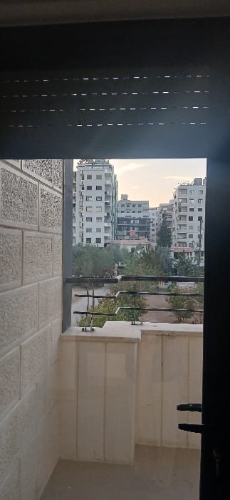 The image depicts a static view from a residential balcony or window in Ashrafia, Syria, looking outwards towards a cityscape under a pale sky. In the foreground, the top portion shows a dark, horizontally slatted blind, partially lowered. To the left, a light-colored, textured block wall with rectangular patterns is visible. A dark metal railing with multiple horizontal bars extends across the mid-foreground, secured to a light-colored concrete ledge. A sliver of a dark vertical frame, possibly a door frame, with a dark handle is seen on the far right. Beyond the balcony, a strip of dense green foliage, consistent with trees and shrubs, separates the foreground from the background. The background features several multi-story residential buildings, primarily light-colored, with numerous windows. One building in the central background displays exposed, darker interior sections on multiple floors, suggesting structural work, renovation, or damage. No individuals, explicit actions, or interactions are visible within the frame. The overall environment appears urban with interspersed greenery.