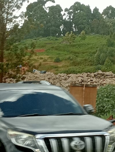 A black Toyota Prado dominates the foreground. Behind the vehicle lies a pile of large rocks, partially obscuring a small, tan-colored structure. Beyond the rocks, a grassy hillside slopes upward towards a dense line of trees. The sky is overcast, casting a muted light over the entire scene. The composition suggests a rural or suburban setting.