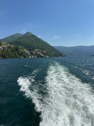The image captures a view from a boat moving on Lake Como near Nesso, Italy. On the left is a shoreline with a steep, densely forested mountain ridge and buildings of Nesso along its base. The right half of the image shows the turbulent wake of the boat, with frothy white water contrasting against the deep blue of the lake. The sky is a clear blue.
