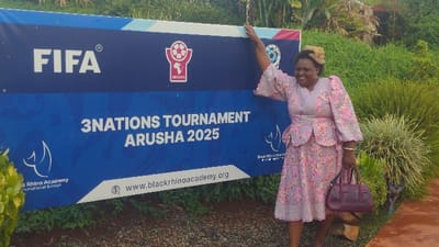 A woman in a pink patterned dress poses next to a large blue sign that reads "FIFA," "3Nations Tournament Arusha 2025," and "Black Rhino Academy." The sign also includes logos for FIFA and CECAFA, along with the website address of Black Rhino Academy. The woman is smiling, with one hand resting on the sign. Lush green foliage fills the background. She carries a burgundy handbag.