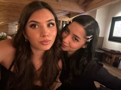 Two women pose closely for a picture. The woman on the left has dark hair adorned with butterfly clips and wears gold hoop earrings. The woman on the right has similar hair, styled in loose waves, and gold hoop earrings. Both women wear dark tops. The background includes a side table, framed art, wooden beams, and flowers. The photo appears to have been taken at an angle.