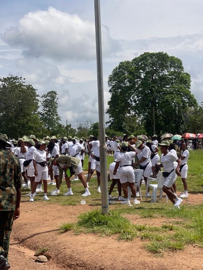 A large group of young adults, predominantly women, are gathered outdoors in a dusty, grassy area under a cloudy sky. They are dressed in white t-shirts and white shorts, many with green and white striped socks. Most are wearing green caps with a national emblem. Some individuals are actively stretching or performing calisthenics, while others stand in clusters, talking. In the foreground, a person in camouflage military uniform is partially visible from the back, facing away from the camera. Behind the participants, there are trees, some market stalls with umbrellas, and a tall metal pole that is a central element in the composition. The scene appears to be a gathering or training event, possibly related to a national youth service program given the inscriptions on a white chair in the background. The location is identified as Orun Ekiti, Nigeria.