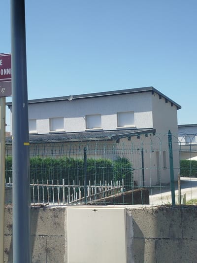 The image captures a street view in the city of Charchilla, France, on a clear, sunny day. The focus is on a modern, two-story building with a light-colored textured facade and a dark, sloping roof. Several windows with white blinds are visible on the upper level. A satellite dish is mounted on the roof. In front of the building, a green metal fence encloses a row of dense green bushes, likely a hedge. Behind the fence, there are some older metal posts with a weathered appearance, interspersed with some darker debris or vegetation. A grey concrete wall forms the foreground, with a white electrical box or utility enclosure situated on top of it. A large grey cylindrical pole, likely a lamppost or utility pole, stands prominently on the left side of the frame, partially obscuring a sign with red and white text, which appears to be a street sign. The sky is a clear, vibrant blue, with no clouds visible. The overall impression is one of a residential or small commercial area under bright daylight conditions.