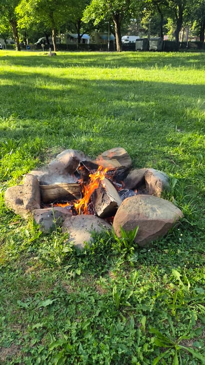 A campfire is burning within a ring of stones on a grassy area. The fire is fueled by logs, with flames and smoke rising from the wood. Several large stones form the perimeter of the fire pit. The surrounding area is covered in lush green grass, with some scattered weeds and small plants. In the background, trees with dense green foliage are visible, and further in the distance, several mobile homes or RVs are parked, suggesting a campground or recreational area. The lighting indicates it is daytime, with sunlight casting shadows on the grass. The location is Pchelishte, Bulgaria.