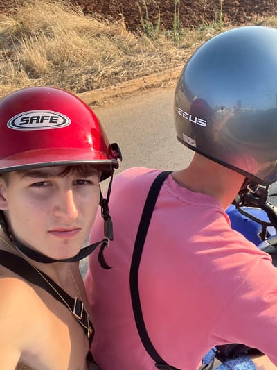 Two people are on a motorcycle. The person in the front is wearing a shiny red helmet with the word "SAFE" on it and a black strap is visible. The person behind is wearing a light grey helmet with the word "ZEUS" on it and a pink shirt with a black strap across the chest. The background shows a road and dry grassy land.