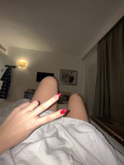 A person in a white robe sits in a room. Their legs are visible, and one hand is raised, displaying a peace sign. The nails are painted with red dots. In the background, there's a wall with several framed pictures, a wall lamp, and a partial view of a bed with patterned sheets. A curtain is visible above the person.