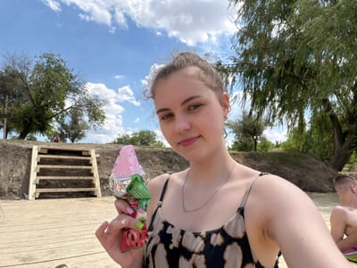 In Novomykolaivka, Ukraine, a young woman, with her hair pulled up in a bun, holds a pink ice cream cone with a wrapper depicting a watermelon slice. She is wearing a patterned tank top and a silver necklace. In the background, wooden stairs lead up a dirt embankment. A man, shirtless, sits to her right. The scene appears to be outdoors on a sunny day with a partly cloudy sky.