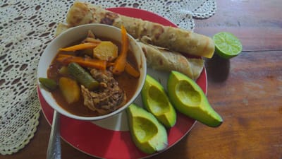 A meal is presented on a dark brown wooden table in an indoor setting in Unknown, Tanzania. A white bowl with a blue rim, centrally positioned on a red plate with white stripes, contains a reddish-brown stew. The stew includes cooked meat, visible chunks of potato, sliced carrots, and green vegetables identified as zucchini or cucumber. To the right, on the same red plate, are three halves of an avocado with pits removed, displaying light green flesh. Also on the plate are several rolled, golden-brown flatbreads and one half of a green lime. A silver-colored spoon with an etched handle is partially visible beneath the bowl. A white crocheted doily with an intricate pattern covers a portion of the table surface in the upper left quadrant. No individuals are present in the scene.