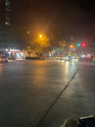The image captures an urban street intersection at night in Dali City, Yunnan Province, China, specifically at or near the intersection of Eryuan Road and Renmin Road. The asphalt road surface in the foreground is wet, exhibiting strong reflections from various light sources. A human hand, partially visible in the bottom right corner, holds a mobile device, indicating the perspective of the photographer.

In the midground, a dark-colored sedan or SUV with illuminated taillights is visible on the left side, appearing to be turning or stopped. Adjacent to it, a single pedestrian stands on the sidewalk. Behind the pedestrian, a multi-story building features illuminated red signage, including characters for "永和豆浆" (Yonghe Doujiang), a prominent Chinese fast-food chain. A bright, yellow-orange streetlight casts a wide glow over the street and several trees. On the right side, multiple vehicles, including several yellow-roofed taxis and at least one motorcycle or scooter with its headlight on, are stationary. They appear to be stopped at a red traffic signal, with a green traffic signal also visible nearby.

The background consists of dark, silhouetted buildings and a dark night sky. The scene is illuminated predominantly by artificial light sources, creating distinct highlights and reflections on the wet pavement.