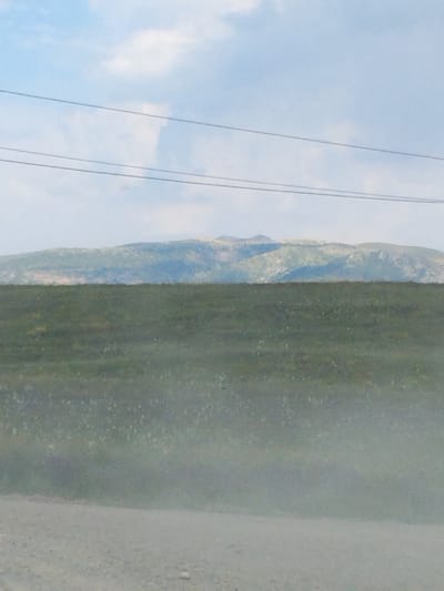 The image depicts a landscape scene featuring a vast expanse of green grassland in the foreground, subtly blurred by atmospheric conditions or a shallow depth of field. In the middle ground, a mountain range with varied textures and colors, ranging from muted greens to earthy browns and hints of rocky outcrops, stretches across the horizon. Above the mountains, a pale blue sky with scattered white clouds is visible. Two thin, dark horizontal lines, presumably power lines, traverse the upper portion of the image. The lower part of the image shows a rough, gravelly surface, suggesting a dirt road or unpaved area. The overall impression is one of a natural, open environment, possibly in or near the city of Ак-Дөбө, Kyrgyzstan, given the context.