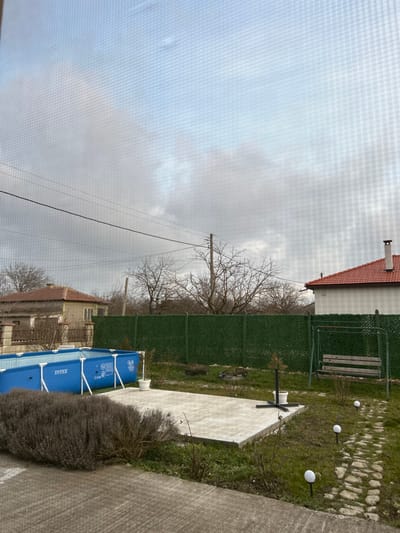 The image depicts an outdoor residential backyard in Tsarichino, Bulgaria, viewed through a fine mesh screen under an overcast, light grey sky. No individuals are present. The foreground features a dense, dark grey shrub occupying the lower left. Centrally, a rectangular light grey concrete patio slab is visible. To the left of this slab, a blue Intex above-ground swimming pool with exposed metal framing stands. A small white potted plant is positioned between the pool and the slab. To the right of the concrete patio, a garden swing set with a wooden bench seat is present. A winding path constructed from irregular light-colored stones extends from the concrete slab towards the right background, lined with three white spherical solar lights on stakes. The ground consists of patchy green grass interspersed with bare soil and small dormant plants. A dark green lattice-patterned fence forms a horizontal barrier across the mid-ground. Beyond the fence, a line of bare deciduous trees indicates a cold season. Utility poles with overhead power lines traverse the upper mid-ground horizontally. In the background, to the left, a light-colored house with a brown tiled roof is partially visible. To the right, another light-colored house with a red tiled roof and a prominent chimney is observed.