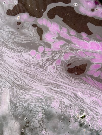 The image displays an abstract, top-down view of a water surface with swirling patterns of brown, grey, and pink. The brown areas appear as larger, irregular masses, while the grey forms fluid, wave-like streaks. Interspersed are clusters of pink, circular shapes. Small, dark specks and occasional lighter-colored bubbles are visible within the grey and pink sections. The textures suggest a foamy or emulsified substance. There are no discernible people, structures, or clear geographical features identifiable in this abstract representation of a liquid surface. The location is stated as Tekeli, Kazakhstan.