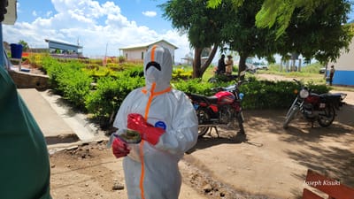 The image displays a person dressed in a full white hazmat suit with bright orange stripes and red gloves, standing on a dirt path in Tchere, Democratic Republic of the Congo. The individual, whose face is obscured by the suit, holds a clear plastic bag containing green items and what appears to be a face shield, with the words "Face Shield" faintly visible. A "Tyvek" logo is visible on the chest of the suit.

The scene unfolds outdoors under a bright blue sky dotted with fluffy white clouds, indicating a sunny midday or early afternoon. In the mid-ground, two motorcycles are parked on the unpaved ground; one is red with "HADJIRA" written on its tank, and the other is a darker maroon. Lush green bushes line a concrete drainage ditch that runs along the path, separating it from a slightly raised area. Several green trees cast shadows, providing shade on the right side of the image.

In the background, a white building with a green roof is visible among other structures, possibly homes or temporary constructions. Faint figures of other people can be seen further back near the buildings and a large water tank structure. The setting appears to be a rural or semi-urban area. The presence of the Personal Protective Equipment (PPE) suggests an ongoing public health activity, such as disease prevention, medical outreach, or response efforts. The photographer's watermark, "Joseph Kisuki," is visible in the bottom right corner.