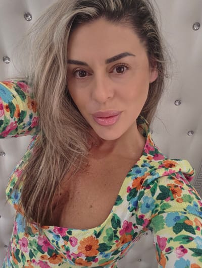 A woman with blonde hair and brown eyes is shown in a close-up, selfie-style shot. She is wearing a yellow floral print top with a low neckline. The background is a white tufted surface with several round, reflective embellishments. The image appears to have been taken indoors. There is no indication of the city of Carpi, Italy, in the image.