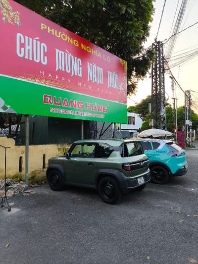 The scene depicts an outdoor urban environment in Phường Nghĩa Lộ, Thành phố Quảng Ngãi, Vietnam. A large, rectangular billboard, primarily red and green, occupies the upper left portion of the image. The red section displays Vietnamese text "PHƯỜNG NGHĨA LỘ CHÚC MỪNG NĂM MỚI" with "HAPPY NEW YEAR" below it, while the green section features "QUANG HÙNG PROFESSIONAL ADVERTISING COMPANY". The billboard is supported by two grey metal posts partially obscuring a low, light-yellow concrete wall to its left.
Two compact SUVs are parked side-by-side on an asphalt surface. The foreground vehicle is an olive green Wuling Mini EV Macaron (or similar model) with black wheels, displaying a Vietnamese license plate "76A-157.15" on its rear bumper. Directly behind it, a light blue/teal VinFast VF 5 Plus SUV is parked, also equipped with black wheels.
To the far left, a black metal garment rack or stand is visible next to the yellow wall, beneath which are stacks of brown and grey patterned tiles. The background features multiple utility poles with dense bundles of overhead wires, green foliage from trees, and distant residential or commercial buildings. A white umbrella or awning structure is discernible behind the light blue vehicle. The sky is bright and overcast, suggesting daylight conditions. No individuals are visible, and no actions or interactions are depicted.