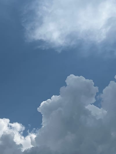 The image shows a clear blue sky with various cloud formations. In the foreground, there are large, fluffy white clouds that appear dense and somewhat shadowed, suggesting they might be developing cumulonimbus clouds. Towards the upper portion of the image, the clouds are lighter and more wispy, indicating cirrus or cumulus clouds at a higher altitude. The sky itself is a vibrant blue, suggesting it is daytime and likely fair weather, despite the presence of these substantial clouds. There are no people, structures, or any elements of a city visible in the frame; the image is exclusively focused on the sky and its cloudscape. The location is Pamyati 13 Bortsov, Russia.