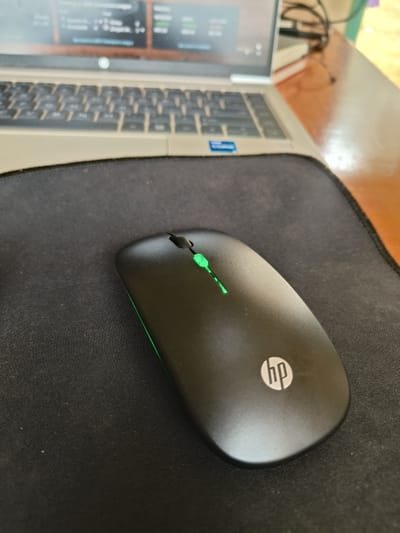 The image displays a close-up, elevated perspective of a computer workstation. In the foreground, a black HP-branded wireless computer mouse with a distinct green LED illumination is centrally positioned on a rectangular black fabric mousepad. The mouse features the HP logo in silver-grey. Behind the mousepad, a silver-grey HP laptop is open, its screen displaying blurred content suggestive of data or graphical interfaces. A blue "Intel Core" sticker is discernible on the laptop's palm rest. Both the laptop and mousepad rest on a polished, light brown wooden desk surface. No human subjects are visible within the frame. This scene is located in the city of Murang'a, Kenya.