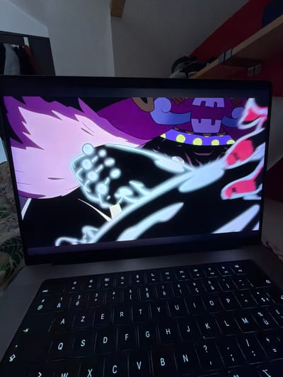 A laptop displaying an animated scene is positioned in a domestic environment. The screen displays a character with purple and white features, alongside bright white animated effects. Next to the screen is the laptop's keyboard, which is backlit. The background reveals partial views of a room with shelving and objects. The image was taken at an interior location; city is not possible to determine from the image. The image does not allow identification of La Salle-les-Alpes.