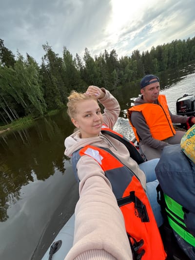 The image shows two people, a woman and a man, in an inflatable boat on a body of water. The woman is in the foreground, taking a selfie. She has blonde hair pulled back and is wearing a light-colored hoodie and an orange life vest. The man is in the background, wearing a baseball cap and an orange life vest, and appears to be operating the boat. The boat is moving, as indicated by the ripples on the water and the spray behind it. The background consists of a dense forest lining the shore of the lake. The sky is overcast with some patches of sunlight. The overall impression is a recreational boat trip on a lake, possibly in a rural or natural area. The location is identified as Anteplevo (Okhotinskoe S/P), Russia.