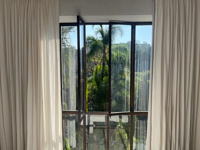 The scene depicts an interior perspective in San Miguel Acapantzingo, Mexico, looking outward through a large, multi-pane window. Two cream-colored, vertically pleated curtains are partially drawn, framing the window on both the left and right sides. The window features dark-toned framing. The upper-left window panel is observed slightly ajar, rotating outward, while the remaining glass panes are closed. Reflections are visible on the window glass surface. Beyond the window, a dense, green tropical landscape is present under a clear blue sky, characterized by numerous palm trees and varied foliage, indicating bright daylight conditions. No individuals or explicit actions are depicted.