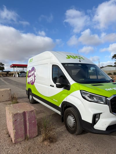 The image depicts a white commercial van, identifiable as a "JUCY" rental vehicle, parked on a gravel and dirt surface at a roadside location in Penong, Australia. The van features a distinctive livery with bright green accents and a purple "Find your Happy!" logo on its side. To the left of the van, two large, weathered, purplish-pink concrete blocks are present on the ground amidst sparse, dry vegetation. In the background, a gas station with a red canopy and multiple fuel pumps is visible. A blue semi-truck with a silver trailer is positioned near the fuel pumps. Further back, low-rise buildings, trees, and scrubland are under a clear blue sky with scattered white clouds. No individuals are visible, and no active interactions are occurring. The scene is illuminated by natural daylight.
