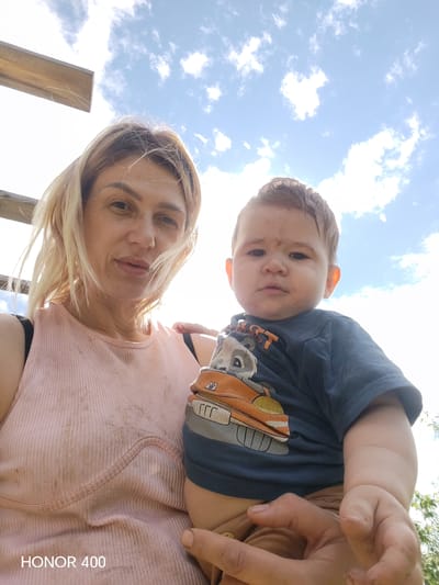 A low-angle, outdoor, selfie-style shot features a woman and a baby against a bright blue sky with scattered white clouds. A wooden plank is visible in the upper left quadrant, and green foliage is visible in the lower right. The woman, in the foreground left, has blonde hair and is wearing a pink ribbed tank top. She is holding the baby, positioned to the right of the frame. The baby has dark hair and is wearing a dark blue t-shirt with a graphic print of a raccoon driving a vehicle. The baby's face has smudges of dirt. The woman's tank top also appears to have dirt stains. The lower left corner of the image displays the text "HONOR 400".