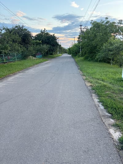 The image depicts a long, straight asphalt road stretching into the distance, bordered by a grassy verge on the left and a wooded area on the right. Overhead power lines run along the left side of the road, supported by utility poles. The sky is partly cloudy with hints of sunset colors, suggesting it is late afternoon or early evening. The weather appears to be clear. The scene is likely rural, given the presence of trees, grass, and the overall quiet atmosphere. There are no people visible in the image. Some green fencing is visible near the edge of the wooded area, and what appear to be small signs attached to the fence. The road itself shows some cracking and wear. The overall mood is one of serenity and a peaceful rural landscape.