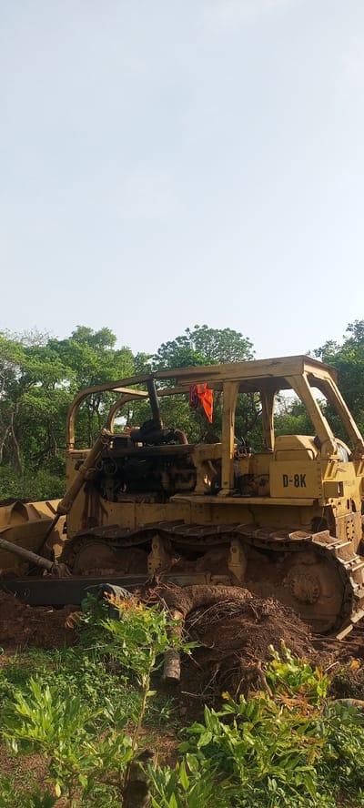 The scene depicts a yellow Caterpillar D-8K bulldozer situated in a vegetated area, possibly on the outskirts of Otunade, Nigeria. The bulldozer appears used, with mud and debris covering its tracks and undercarriage. An orange cloth is draped inside the roll cage of the machine. The surrounding environment consists of green foliage, including bushes and trees, suggesting a rural or undeveloped setting. The sky is a pale blue, indicating daylight. No people are visible in the image. The bulldozer appears to have been operating recently, given the disturbed earth around it.