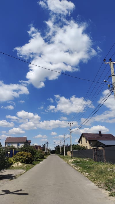 This is a vertical, eye-level, medium-shot photograph taken on a sunny day in the city of Aidarovo, Russia. The scene depicts a paved, empty road stretching into the distance, flanked by residential houses and vegetation. The sky is a vibrant blue, dotted with fluffy white clouds. Overhead, power lines are strung between utility poles, casting shadows on the road. On the left side of the road, a house with a brown roof and white walls is visible, surrounded by lush green bushes and trees. On the right, another house with a brown roof and yellow brick walls is seen behind a dark fence. The overall impression is a peaceful, suburban street scene on a bright summer day.