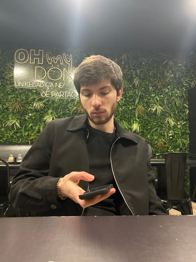 A man with brown hair and a beard sits at a table, wearing a black jacket over a black sweater. He holds a black smartphone in his right hand. Behind him is a wall covered in greenery, with a neon sign that reads "OH my DON" and "UN KEBAB CA NE SE PARTAGE." A chair can be seen in the background, as well as a part of the table on the right. The man appears to be looking down at his phone.