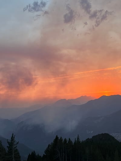 This image captures a dramatic mountain sunset over San Juan de Plan, Spain. The sky is ablaze with vibrant orange and pink hues, illuminating streaks of clouds and casting a warm glow over the landscape. Silhouetted against the fiery sky are the majestic peaks of the Pyrenees, their rugged outlines softened by the fading light. A dense forest of pine trees occupies the right side of the frame, their dark forms providing a stark contrast to the colorful sky.

The scene evokes a sense of awe and tranquility, as the day transitions into night. The air appears clear, with no indication of adverse weather, suggesting a peaceful evening. The rotation of the image suggests it was taken in a vertical orientation, likely to capture the full expanse of the sky and mountains. The presence of the mountain range and coniferous forest strongly suggests a location in a mountainous region, consistent with San Juan de Plan in the Spanish Pyrenees. No people or specific objects are visible, focusing the viewer's attention entirely on the natural beauty of the sunset and the mountain landscape. The time of day is clearly evening, specifically sunset, as evidenced by the intense colors in the sky.