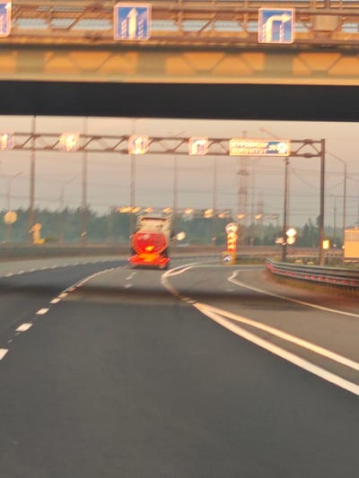 An outdoor, medium shot captures a road scene at dawn or dusk in Torfyanoye, Russia. The sky is hazy and illuminated with warm, orange light, suggesting early morning or late evening. The road ahead is paved and marked with white lines. In the foreground, the road curves to the right. A silver tanker truck with red taillights is visible in the middle distance, traveling away from the camera. Overhead, a road sign gantry is present with multiple illuminated signs. One sign displays text in Cyrillic and English: "КУРОРТНЫЙ 28 KUROVITSY 28". Another sign shows a right-turn arrow. To the right of the road, a metal guardrail with red reflectors lines the edge. The background features a line of trees and utility poles. The overall impression is one of a quiet highway during low light conditions.