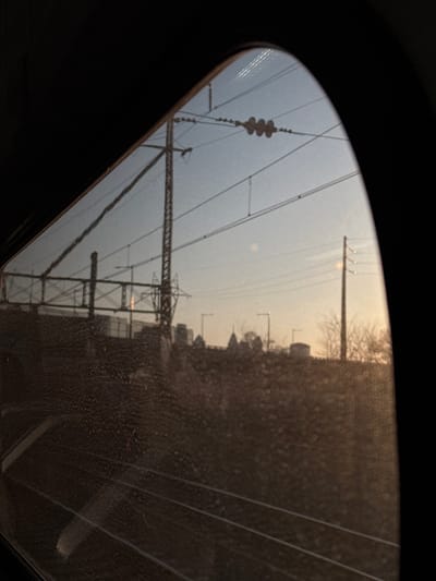 The image displays an exterior urban landscape viewed through a dirty, curved window, likely from a train or subway car, characterized by a dark, curved frame. The window glass is obscured by numerous water droplets or smudges, and subtle horizontal lines suggesting train tracks are visible through the lower portion of the dirty glass.

The sky in the background is a pale blue, transitioning to a golden-orange hue at the horizon, indicating either sunrise or sunset. Dominating the upper left and mid-ground are elements of a railway catenary system, including multiple overhead electrical wires and a tall, dark metal lattice tower. To the right of the lattice tower, an elevated structure or bridge with a metal railing is visible.

Below the infrastructure, a raised landmass or embankment is present, sparsely covered with bare trees or bushes, consistent with a dormant season. Several streetlights are spaced along this embankment. In the distant urban skyline, various buildings are silhouetted against the horizon. A distinctive building featuring a central dome-like structure flanked by two smaller turrets is prominently visible. This specific architectural silhouette identifies the distant landmark as the **Reading Terminal Headhouse**, which is part of the Pennsylvania Convention Center.

The scene is located in **Philadelphia, Pennsylvania, United States of America**. The view is from an elevated train track, likely an approach to **30th Street Station**, looking generally **eastward towards Center City**. No people or direct interactions are visible.