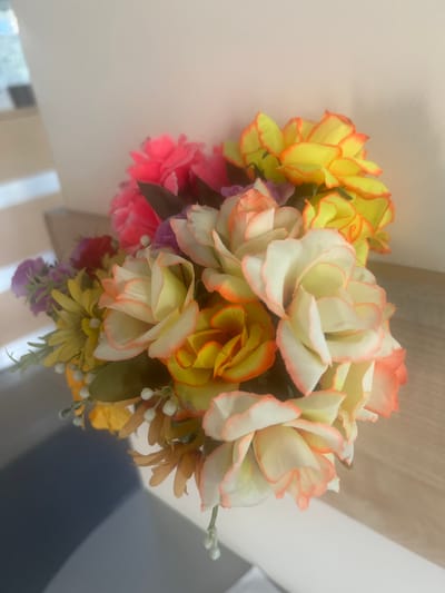 A close-up, slightly out-of-focus shot shows a vibrant bouquet of artificial flowers. The flowers are in various stages of bloom, with prominent shades of yellow, orange, white, pink, and purple. Some of the flowers have delicate pink or orange-tinged edges. Small white buds and green foliage are interspersed throughout the arrangement. The bouquet is positioned on a light-colored, wooden surface, likely a shelf or table, with a light-colored wall in the background. A subtle shadow of the bouquet is cast on the lower left of the frame, suggesting a light source from the right. The environment appears to be indoors, possibly a home or office setting. There are no people or animals present in the image. The specific location within the city of Magas, Russia, cannot be determined from the visual information provided.