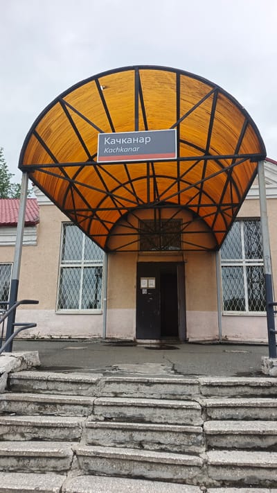 The image shows the entrance to a building with a prominent orange, semi-circular canopy supported by a black metal frame. A sign is affixed to the canopy, displaying the name "Качканар" in Cyrillic script and "Kachkanar" in Latin script. The building facade is a light beige color with two large windows on either side of the entrance. The windows have white frames and are covered with decorative metal grilles. A dark-colored double door is visible under the canopy, with one door slightly ajar. Two small signs are posted to the left of the door. In front of the entrance, a wide set of concrete stairs with visible cracks and wear leads down to an asphalt area. The sky is overcast. The overall scene suggests a public building, possibly a train station or administrative office, in the city of Kachkanar, Russia, given the signage.