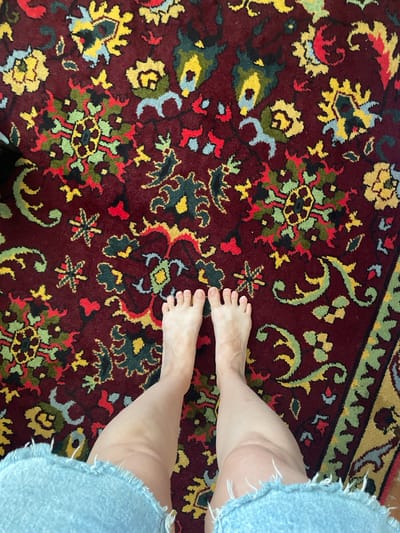The image shows a person's bare feet and lower legs standing on a burgundy rug with a floral pattern. The rug features intricate designs in yellow, green, red, and blue. The person is wearing light blue denim shorts with frayed edges. The scene is photographed from a high angle, looking down at the person's feet and the rug. The location is not specified in the image, and there is no visual information to indicate it is in Kopeysk, Russia.