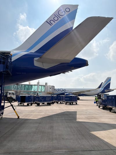 An Indigo Airlines airplane is parked on the tarmac at an airport in Vikram Nagar, India. The tail of the airplane, with the Indigo logo, is visible. Several baggage carts are lined up on the tarmac, some with the Indigo logo and "Ground Force" printed on them. A worker in a yellow vest is walking near the carts. In the background, another Indigo Airlines plane is visible, along with a jet bridge and a ground vehicle. The sky is blue with some clouds.