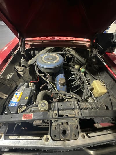 The image shows the open hood of a red vehicle, revealing the engine compartment. Visible components include the engine itself with a blue air filter and valve cover, a black battery labeled "Moura," the radiator, various hoses and wires, and a translucent yellow fluid reservoir. The vehicle appears to be older, based on the engine design. The location is not discernible from the image itself. There is no verifiable information regarding the city of Munro, Argentina.